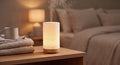A cylindrical LED diffuser lamp with a textured white body and a light wood base, emitting a warm glow and mist, placed on a wooden bedside table in a cozy bedroom setting.